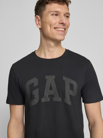 Männermodel trägt T-Shirt mit Logo und Rundhalsausschnitt von GAP in Schwarz, Reine Baumwolle, Regular-Fit, Logo-Print, Nahaufnahme Oberkörper. Rückenlänge bei Größe M 72 cm.
