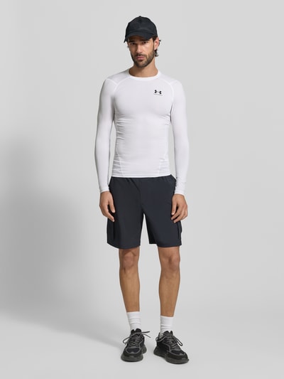 Mannelijk model draagt Slim Fit Longsleeve met Logo-Print van Under Armour, elastische materiaalmix, slim fit, logoprint, ronde hals, zwarte korte broek, volledig lichaam vooraanzicht.