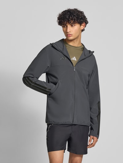 Męski model nosi Slim Fit Funktionsjacke aus schnelltrocknendem Material von adidas Sportswear, efekt: jednolity kolor, krój slim fit, kształt kołnierza: kaptur, zapięcie: zamek błyskawiczny, aplikacja: nadruk z logo, paski w kontrastowym kolorze, rękawy: długi rękaw, funkcja: szybkoschnący, regulujący pot, boczna kieszeń zapinana na zamek błyskawiczny. Ganzkörperansicht z przodu.