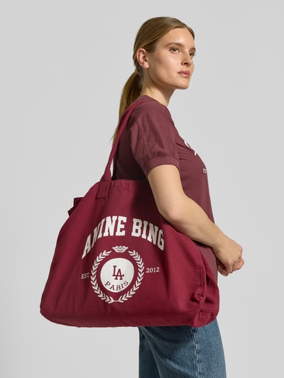 Vrouwenmodel draagt Shopper uit pure katoen van Anine Bing in bordeauxrood met labelprint, hoofdvak open, smalle schouderriemen, canvasstructuur, voorkant zijaanzicht, Shopper uit reiner Baumwolle van Anine Bing, breedte 60 cm, lengte 46 cm, diepte 15 cm.