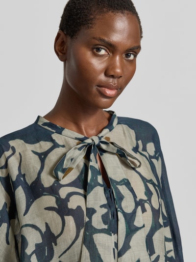 Kobiece model prezentuje bluzka z kokardą i rękawami z falbanami 'Bluse mit Schluppe und Volantärmeln' od Luisa Cerano, mieszanka lyocellu z dodatkiem ramii, krój loose fit, wycięcie w kształcie łezki, szerokie rękawy z marszczonym detalem, długość tyłu w rozmiarze 36: 74 cm, długość rękawów w rozmiarze 36: 65 cm, wyrazisty wzór, odcienie zieleni i beżu, widoczna tekstura tkaniny, zbliżenie górnej części ciała.
