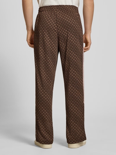 Mężczyznamodel nosi spodnie dresowe Loose Fit Sweatpants mit Logo-Muster model 'FIREBIRD' od adidas Originals, efekt: wzór z logo na całej powierzchni, krój loose fit, wyhaftowane logo, elastyczny ściągacz z tunelem, boczne kieszenie zapinane na zamek błyskawiczny, wykończenie materiału: dzianina dresowa, całe spodnie w brązowych odcieniach, tekstura miękka, tło neutralne, Ganzkörperansicht Rückansicht.