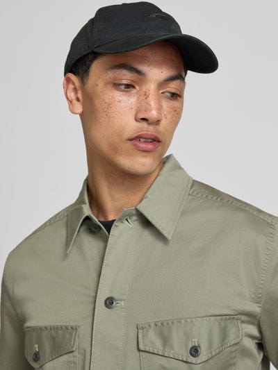 Męski model nosi Hemdjacke mit Kentkragen und Brusttaschen von G-Star Raw, odcień zielony, krój regular fit, kołnierzyk typu kent, listwa guzikowa, kieszenie na piersi, długość tyłu w rozmiarze M 45 cm, szerokość ramion w rozmiarze M 61 cm, szerokość w rozmiarze M 58 cm, długość rękawów w rozmiarze M 67 cm, tekstura gładka, zbliżenie na górną część torsu