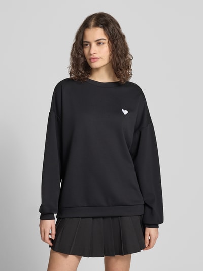 Frauenmodel trägt unifarbenen schwarzen Relaxed Fit Pullover mit Herz-Stickerei von Pieces, Rundhalsausschnitt, Ganzkörperansicht von vorne.