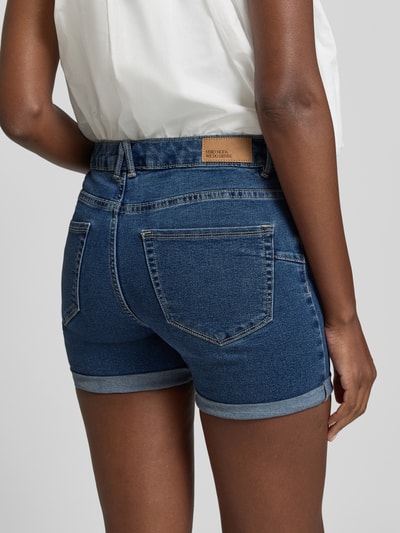 Frauenmodel trägt Regular Fit Jeansshorts aus Baumwoll-Mix Modell 'LUNA' von Vero Moda in Denim-Optik mit Label-Patch, 5-Pocket-Design und fixiertem Umschlag, Ganzkörperansicht Rückansicht.