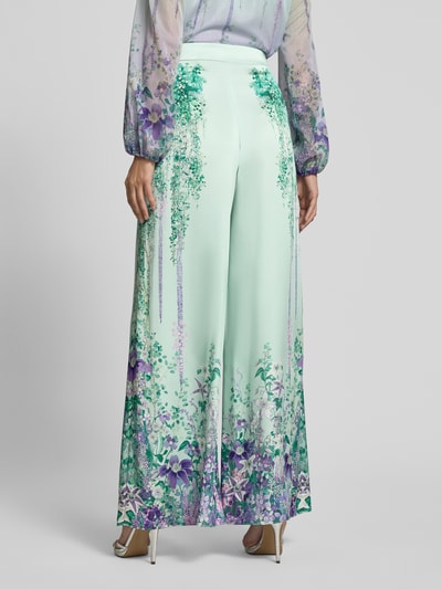 Frauenmodel trägt Hose aus Satin mit weitem Bein von Marc Cain, grün mit Allover-Look, florale Muster, seitlicher Reißverschluss, Ganzkörperansicht Rückansicht.