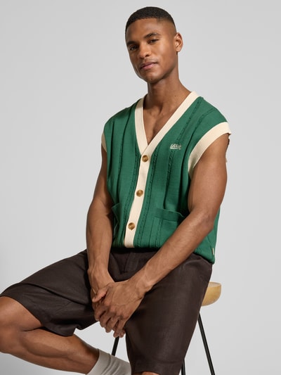 Mannelijk model draagt Regular Fit Weste in breilook met logo-stitching van BLKVIS, groen met beige, opgestikte zakken, v-hals, knoopsluiting, regular fit, vooral groentinten, textuur met strepen, detail van logo, zichtbare knopen, zichtbare naden, zijaanzicht bovenlichaam