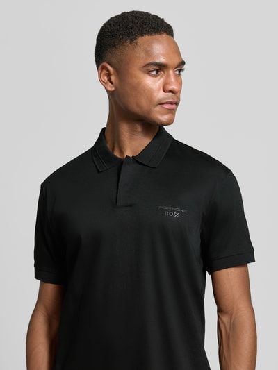 Männermodel trägt Regular Fit Poloshirt aus reiner Baumwolle Modell 'PS Spirit 70 Plk' von BOSS Green X PORSCHE in Schwarz, Nahaufnahme Oberkörper.