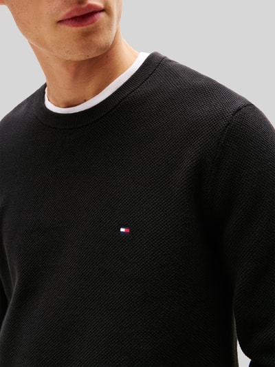 Männermodel trägt Regular Fit Strickpullover aus reiner Baumwolle von Tommy Hilfiger, schwarz, Strukturmuster, Logo-Detail, Nahaufnahme Oberkörper.