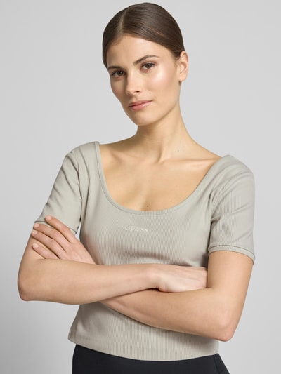 Kobiece modelka nosi Cropped T-Shirt aus Lyocell-Mix Modell 'Circe' od Guess Activewear, jasnoszary, prążkowana tekstura, krój slim fit, logo na piersi, dekolt okrągły, mieszanka lyocellu z bawełną, krótka długość, zbliżenie na górną część ciała.