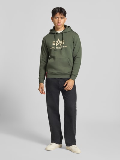 Männermodel trägt Regular Fit Hoodie aus Baumwoll-Mix mit Logo-Print von Alpha Industries in Olivgrün, elastische Abschlüsse, Ganzkörperansicht von vorne.