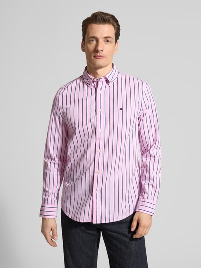 Męski model nosi Regular Fit Freizeithemd aus reiner Baumwolle von Tommy Hilfiger, różowy z granatowymi paskami, długi rękaw, bawełna, krój regular fit, popelina, guzik, logo, widok całej sylwetki z przodu.