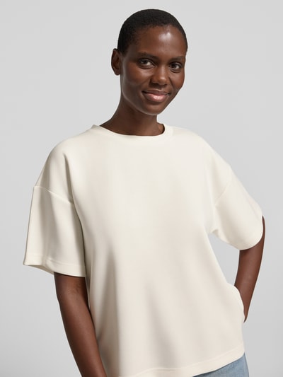 Modelka kobieca ma na sobie damski t-shirt z mieszanki materiałów z dodatkiem wiskozy, pudełkowy krój, model 'Helle', okrągły dekolt, długość tyłu 60 cm w rozmiarze XS/S od My Essential Wardrobe, jasny kolor, gładka faktura, lekko opuszczone ramiona, zbliżenie górnej części ciała z przodu.