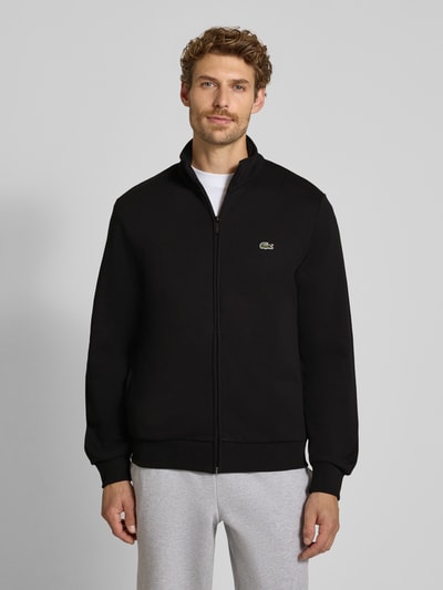Männermodel trägt schwarze Regular Fit Sweatjacke aus Baumwoll-Mix von Lacoste, helle Hose, schlichter Hintergrund, Ganzkörperansicht von vorne. Herren Sweatjacke von Lacoste.