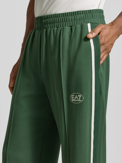 Männermodel trägt grüne Trackpants aus Viskose-Mix von EA7 Emporio Armani mit Galonstreifen und Label-Print, Nahaufnahme Oberkörper.