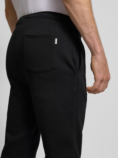 Mężczyzna nosi spodnie dresowe Sweatpants mit Eingrifftaschen Modell 'CERES' von ONLY & SONS, mieszanka bawełny, wpuszczane kieszenie, kieszeń z tyłu, prosty krój, elastyczny pas z tunelem, elastyczne zakończenia nogawek, detal czarny, tekstura gładka, detailaufnahme tylnej kieszeni.