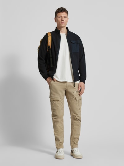 Męski model nosi Sweatjacke mit Stehkragen und Brusttasche von camel active, czarny kolor, krój regular fit, kieszeń na piersi, kieszenie z zamkiem, długość tyłu 68 cm, długość rękawa 67 cm, widok całej sylwetki z przodu.