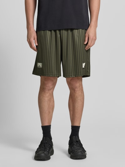 Männermodel trägt Regular Fit Shorts mit Label-Print Modell 'Lama' von Wood Wood in Olivgrün mit dünnen schwarzen Streifenmuster, Ganzkörperansicht von vorne. Elastischer Bund sichtbar, strukturierte Textur, Kontext: Herren Shorts, Bundweite 40 cm, Innenbeinlänge 19 cm.