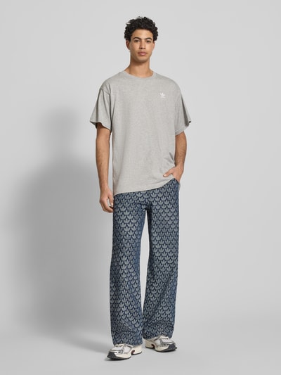 Männermodel trägt T-Shirt mit Label-Stitching von adidas Originals in Grau, reine Baumwolle, Regular Fit, Ganzkörperansicht von vorne.