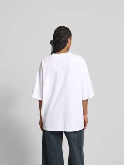 Frauenmodel trägt Oversized T-Shirt mit Label-Stitching von black palms aus reiner Baumwolle, halbärmelig, weiße Farbe, Rückenlänge 70 cm, Ganzkörperansicht Rückansicht.