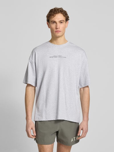 Männermodel trägt T-Shirt mit Label-Print und Rundhalsausschnitt von HALO in Hellgrau, Boxy Fit, reine Baumwolle, Ganzkörperansicht von vorne.
