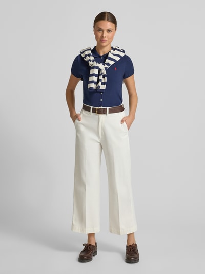 Frauenmodel trägt Poloshirt mit Knopfleiste von Polo Ralph Lauren, dunkelblau, Baumwoll-Elasthan-Mix, Slim Fit, Hemdblusenkragen, Label-Stitching, kombiniert mit weißen weiten Hosen und braunem Gürtel, Rückenlänge bei Größe S 55 cm, Ganzkörperansicht von vorne.