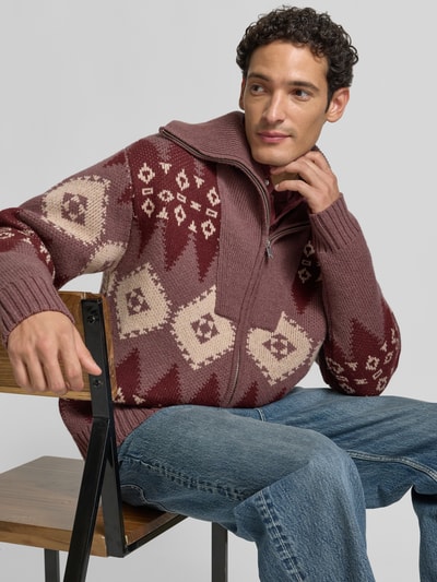 Männermodel trägt Strickjacke mit abstraktem Muster von DRYKORN in Braun- und Beigetönen, Baumwolle, Reißverschluss, Nahaufnahme Oberkörper.