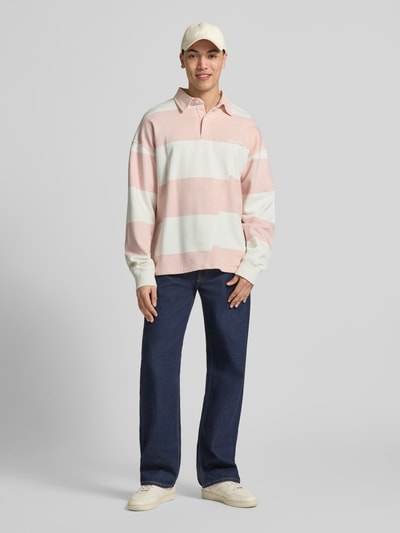 Mężczyzna nosi Relaxed Fit Poloshirt aus reiner Baumwolle Modell 'Castelluccio' von Pegador w pastelowych różowych i białych pasach, bawełna, krój relaxed fit, kołnierzyk polo z zapięciem na guziki, ściągacze na rękawach, haft z logo, obniżone ramiona, długość tyłu w rozmiarze M: 69 cm, widok całej sylwetki z przodu.