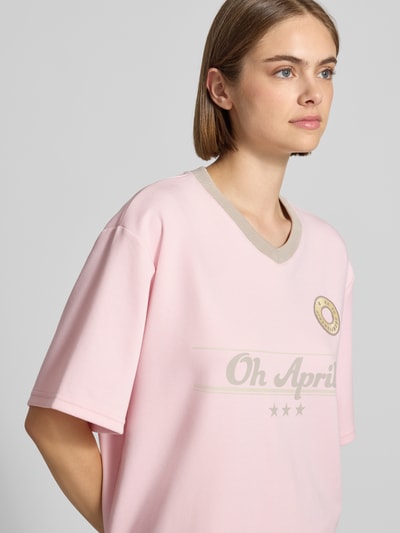 Frauenmodel trägt Trikot mit Label-Print und V-Ausschnitt von OH APRIL in Rosa, Relaxed Fit, sichtbares Stitching, Nahaufnahme Oberkörper.