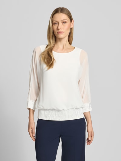 Kobiece modelka nosi Blusenshirt mit Ziersteinbesatz od Vera Mont, krój regular fit, okrągły dekolt, rękaw o dł. 3/4, długość tyłu w rozmiarze 36: 59 cm, białe z ozdobnymi kamieniami, gładka tkanina. Ganzkörperansicht von vorne.