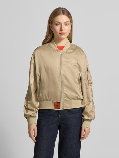 Vrouwenmodel draagt Bomberjacke met Reißverschlusstasche am Ärmel van Original Bombers in beige met opstaande kraag, ritssluiting, gestikt logo, zij-steekzakken met drukknoopsluiting, labelpatch, elastische boorden; ruglengte maat S: 49 cm, schouderbreedte: 50 cm, borstwijdte: 58 cm, mouwlengte: 61 cm; los model. Ganzlichaamsvoorzijde.