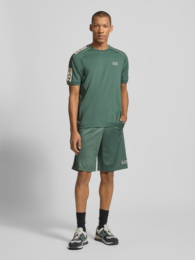 Męski model trägt Szorty mit Logo und wpuszczane kieszenie von EA7 Emporio Armani, zielony kolor, krój relaxed fit, szerokość w rozmiarze M 42 cm, długość wewnętrzna nogawki 28 cm, Ganzkörperansicht von vorne.