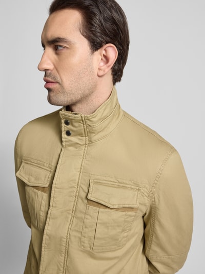 Męski model nosi Jacke mit Stehkragen und Pattentaschen od Christian Berg Men, bawełna z elastanem, krój regular fit, kolor beżowy, z widocznymi patkami na kieszeniach, szerokość w rozmiarze M: 56 cm, długość tyłu: 73 cm, szerokość ramion: 43 cm, długość rękawa: 67 cm, zbliżenie na górną część tułowia, widok z przodu.