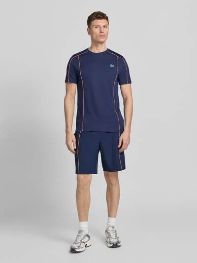 Männermodel trägt Regular Fit T-Shirt mit UV-Schutz 50+ von Lacoste Sport in Dunkelblau, unifarben mit Kontraststreifen, Logo-Patch und Logo-Print, schnelltrocknendes Piqué-Material, Ganzkörperansicht von vorne.