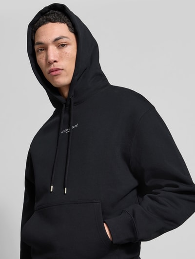 Männermodel trägt Hoodie mit Label-Stitching von MAISON KITSUNÉ in Schwarz, lockere Passform, Kapuze, Kängurutasche, Nahaufnahme Oberkörper.