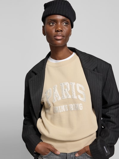 Frauenmodel trägt Sweatshirt aus reiner Baumwolle von Anine Bing in Beige mit Label-Detail, lockere Passform, Rundhalsausschnitt, elastische Abschlüsse, Ganzkörperansicht von vorne.