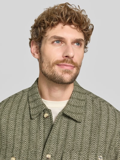 Mężczyznamodel nosi Relaxed Fit Hemdjacke mit Kentkragen und Brusttaschen od Scotch & Soda, mieszanka materiałów z dodatkiem wełny, wzór na całej powierzchni, detal z logo, zielone odcienie, tekstura skośnych linii, zbliżenie na górną część ciała.