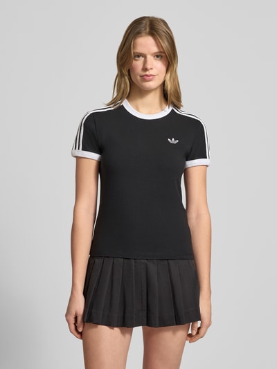 Kobiece modelka nosi Slim Fit T-Shirt aus Baumwoll-Mix od adidas Originals, efekt: jednolity kolor, krój regular fit, wyhaftowane logo, paski w kontrastowym kolorze, okrągły dekolt, krótki rękaw, mieszanka bawełny; widok całej sylwetki z przodu.