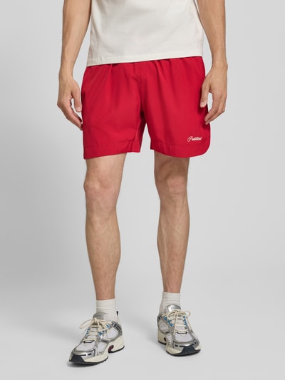 Männermodel trägt Badehose mit Label-Stitching und elastischem Bund von PROHIBITED in leuchtendem Rot, Regular-Fit, Ganzkörperansicht von vorne.