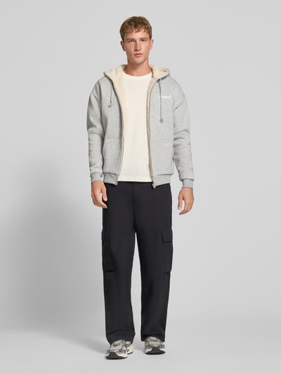 Männermodel trägt Regular Fit Sweatjacke aus Baumwoll-Mix mit Teddyfutter von REVIEW, graue Farbe, Logo-Detail, Ganzkörperansicht von vorne.