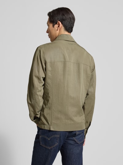Männermodel trägt Regular Fit Jacke aus Lyocell-Mix von Guess in Olivgrün, elastischer Bund, glatte Struktur, Ganzkörperansicht Rückansicht. Rückenlänge 72 cm, Schulterbreite 56 cm, Brustweite 58 cm, Eingrifftaschen mit Reißverschluss.
