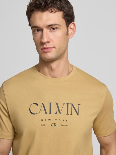 Männermodel trägt T-Shirt mit Label-Print und Rundhalsausschnitt von CK Calvin Klein in Beige, Reine Baumwolle, Regular-Fit, Nahaufnahme Oberkörper.