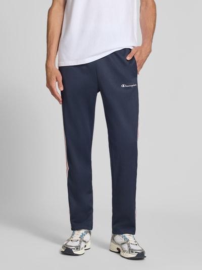 Männermodel trägt Sweatpants mit Bordüre von Champion in Dunkelblau, Material-Mix, gerader Schnitt, Seitentaschen, Nahaufnahme Unterkörper.