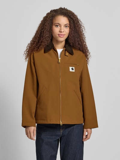 Frauenmodel trägt Jacke mit Stehkragen und Zweiwege-Reißverschluss Modell 'Clark' von Carhartt Work In Progress, krój relaxed fit, detal z logo, długość tyłu w rozmiarze S: 66 cm, szerokość ramion w rozmiarze S: 50 cm, szerokość klatki piersiowej w rozmiarze S: 54 cm, długość rękawów w rozmiarze S: 58 cm, kolor brązowy, gładka faktura, Ganzkörperansicht von vorne.