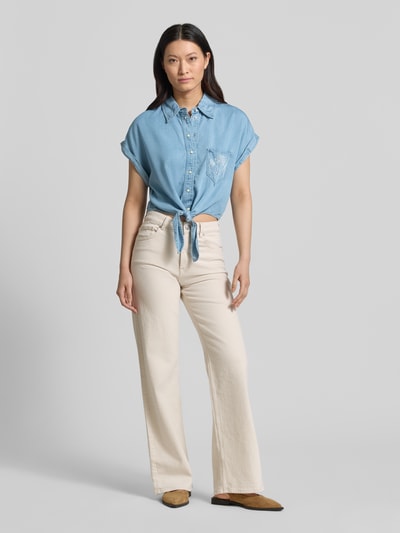 Modelka kobieca nosi bluzkę jeansową z lyocellu, krój loose fit, model 'AMBRA' od Guess, błękit, kieszeń z haftem, wiązany detal, krótki rękaw, listwa guzikowa, kołnierzyk typu kent, długość tyłu 47 cm, długość rękawów 8 cm, widok całej sylwetki z przodu