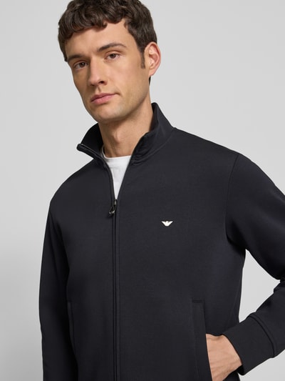 Männermodel trägt Regular Fit Sweatjacke mit Logo-Stitching von Emporio Armani, dunkler Baumwoll-Mix, Stehkragen, Zweiwege-Reißverschluss, Eingrifftaschen, elastische Abschlüsse, Nahaufnahme Oberkörper.