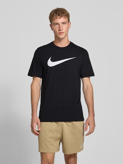 Männermodel trägt Regular Fit T-Shirt aus reiner Baumwolle von Nike in Schwarz mit weißem Logo, beige Shorts, Ganzkörperansicht von vorne.