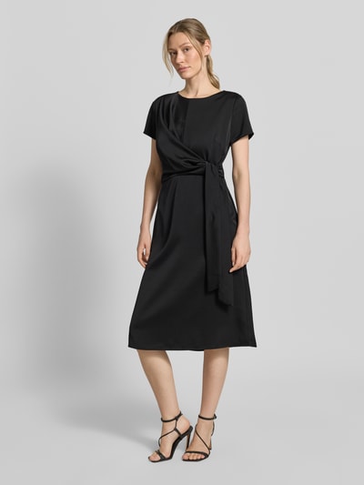 Frauenmodel trägt Knielanges Cocktailkleid mit Knotendetail und Reißverschluss von Rinascimento, schwarze Farbe, Taillierter Schnitt, Rundhalsausschnitt, Knielänge, Rückseitiger Reißverschluss, Rückenlänge 105 cm, Brustweite 46 cm, Ärmellänge 17 cm, Ganzkörperansicht von vorne.