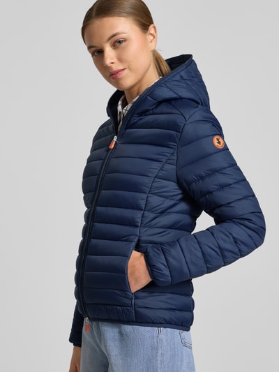 Frauenmodel trägt dunkelblaue Steppjacke mit Kapuze Modell 'DAISY' von SAVE THE DUCK, körpernahe Passform, Ganzkörperansicht von vorne.