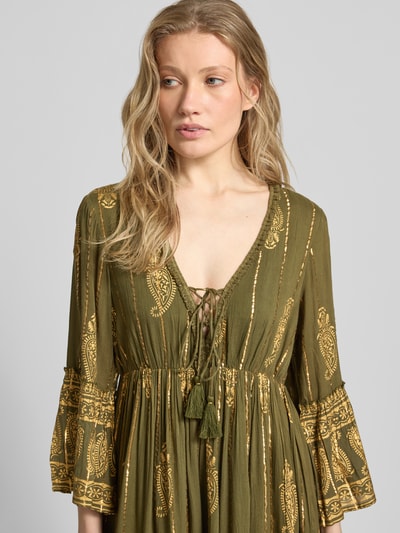 Frauenmodel trägt Minikleid aus Viskose-Mix mit Schnürung von Apricot in Oliv mit goldenen Mustern, Trompetenärmel, Nahaufnahme Oberkörper. Rückenlänge bei Größe S: 83 cm, Brustweite: 35 cm, Ärmellänge: 50 cm, A-Linie, V-Ausschnitt.